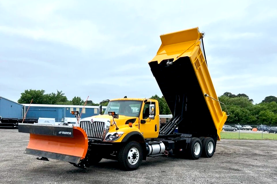 Yellow dump truck with its bed raised, parked on a gravel lot, with trees and a fence in the background.