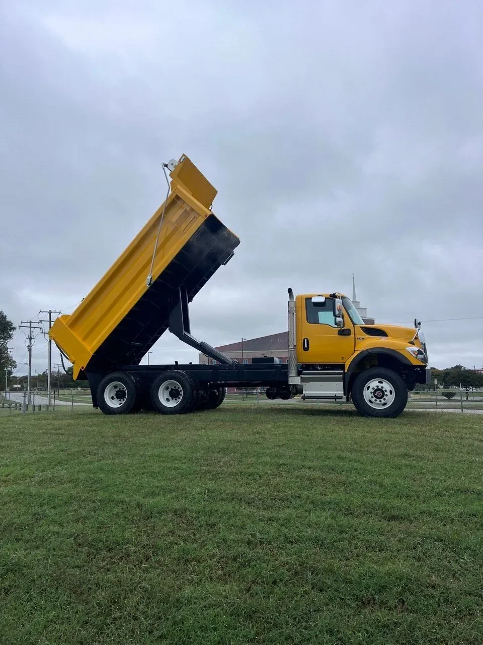 Yellow dump truck with raised bed on grassy area, overcast sky, power lines in background.