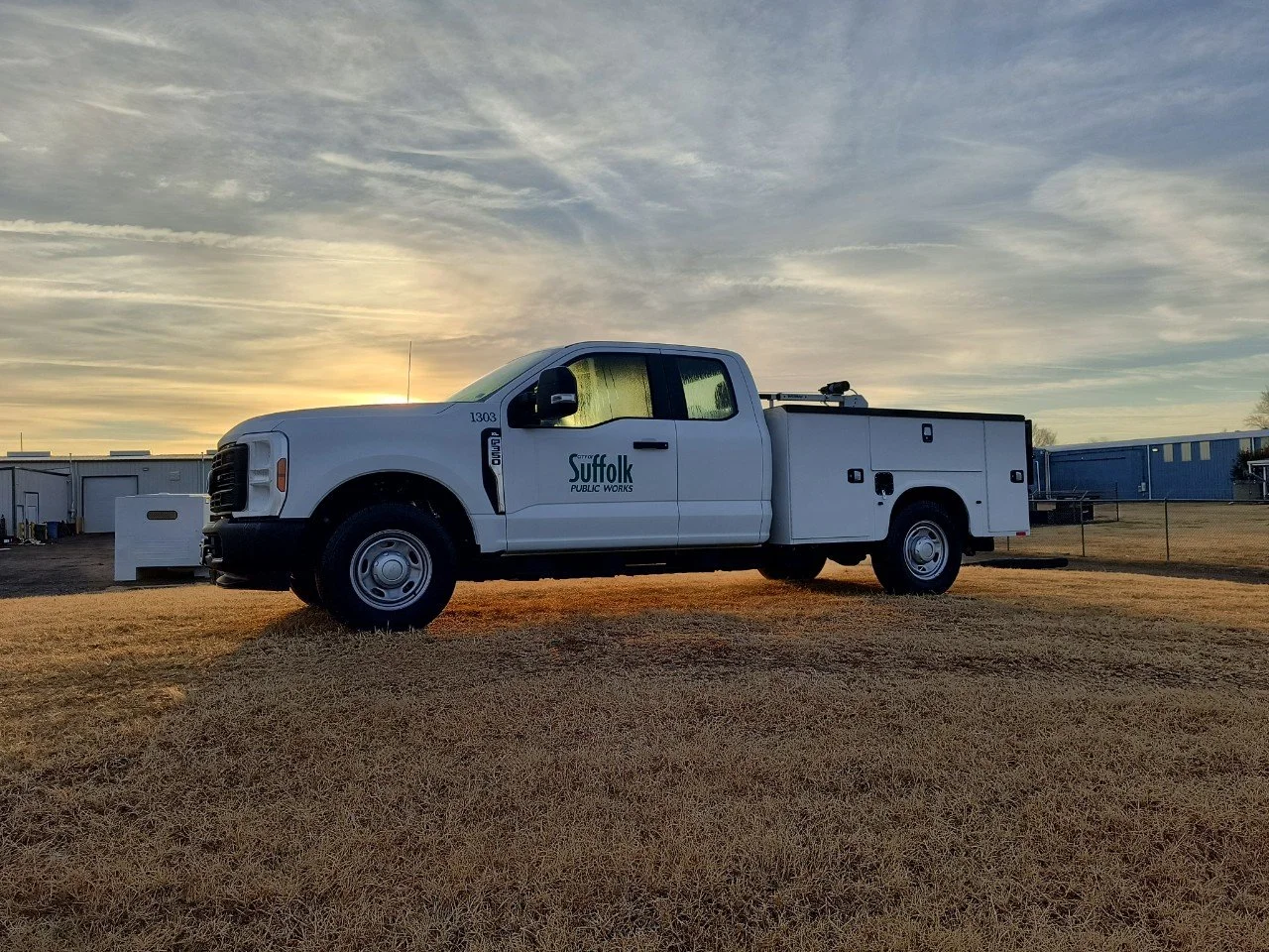 White Suffolk Public Works truck parked on a grassy field at sunset, with buildings in the background.