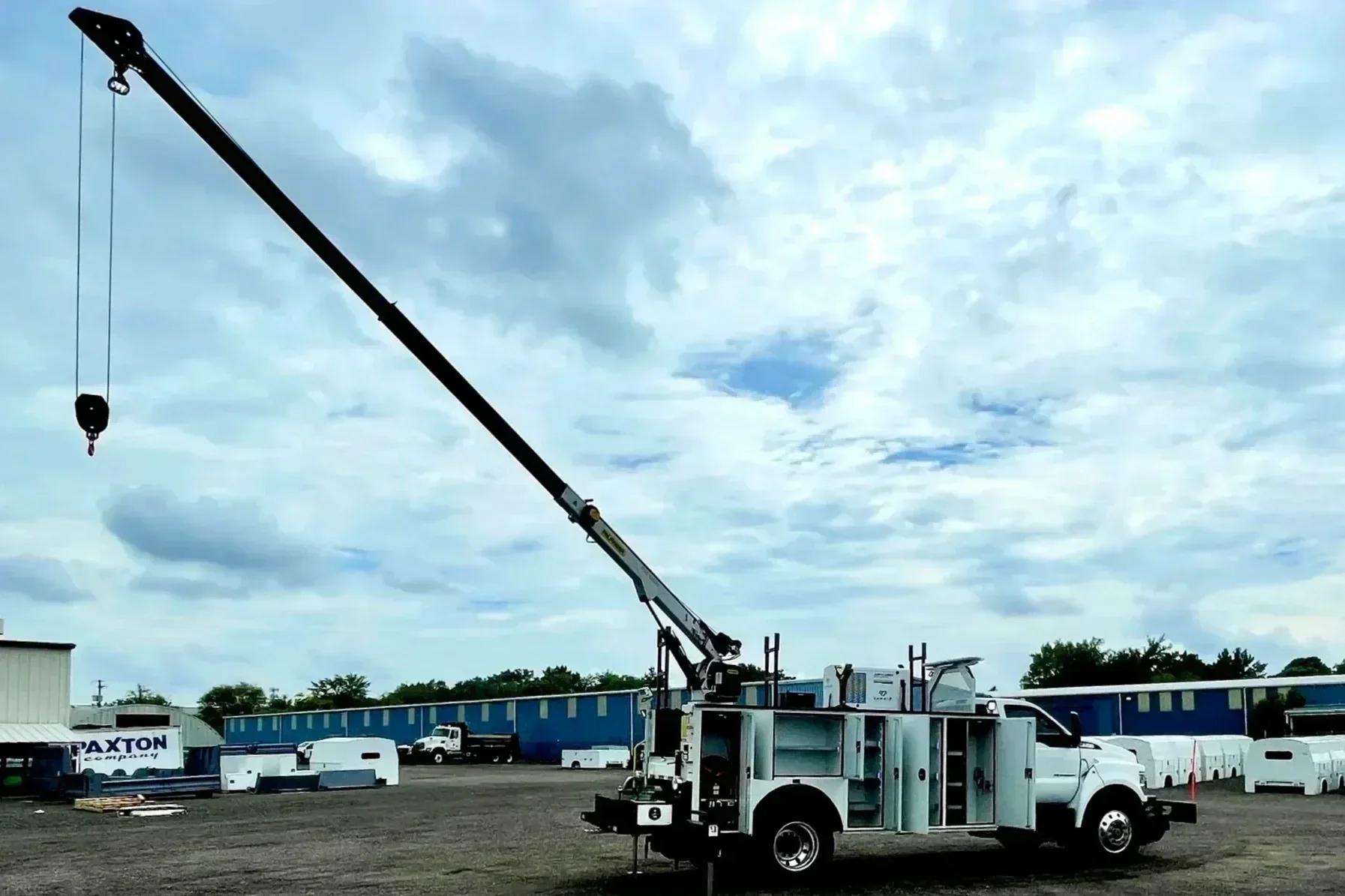 A utility truck with a mounted crane extended into the sky, set in a commercial lot with storage buildings, parked trucks, and cloudy sky.