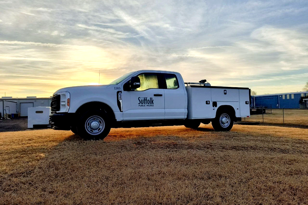 A white Suffolk Public Works utility truck parked on a grassy field at sunset, with buildings and a cloudy sky in the background.