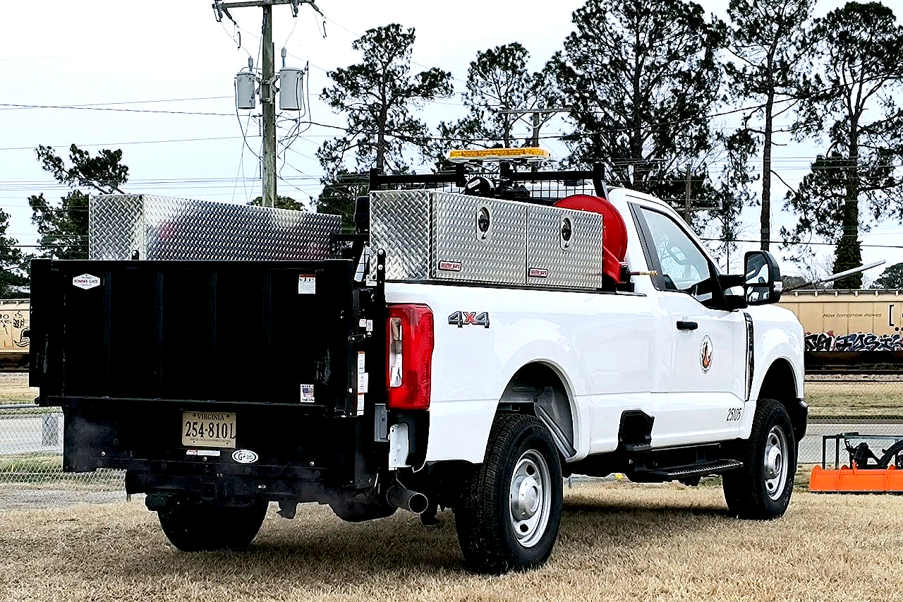 White utility truck with equipment on the back, parked on a grassy area under trees, with power lines in the background.