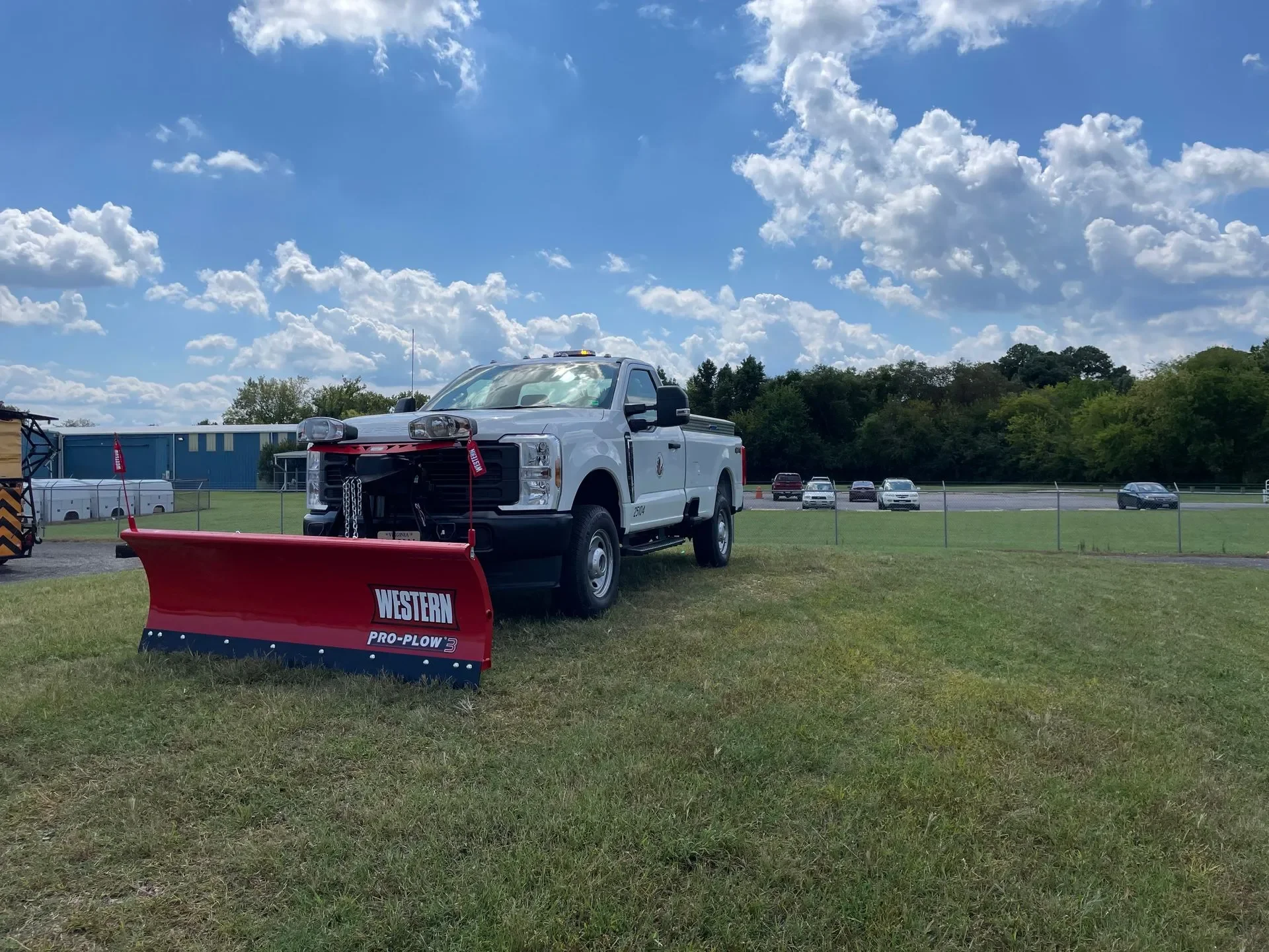 A white pickup truck with a yellow light on top is parked on grass next to a red Western PRO-PLOW 3 snow plow blade. In the background, there are several parked cars, a blue building, and trees under a partly cloudy sky.