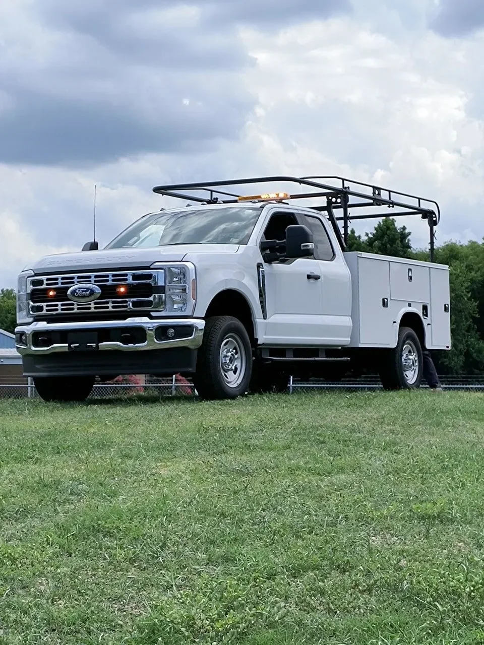 White Ford utility truck with a service body and ladder rack on grass, under cloudy sky.