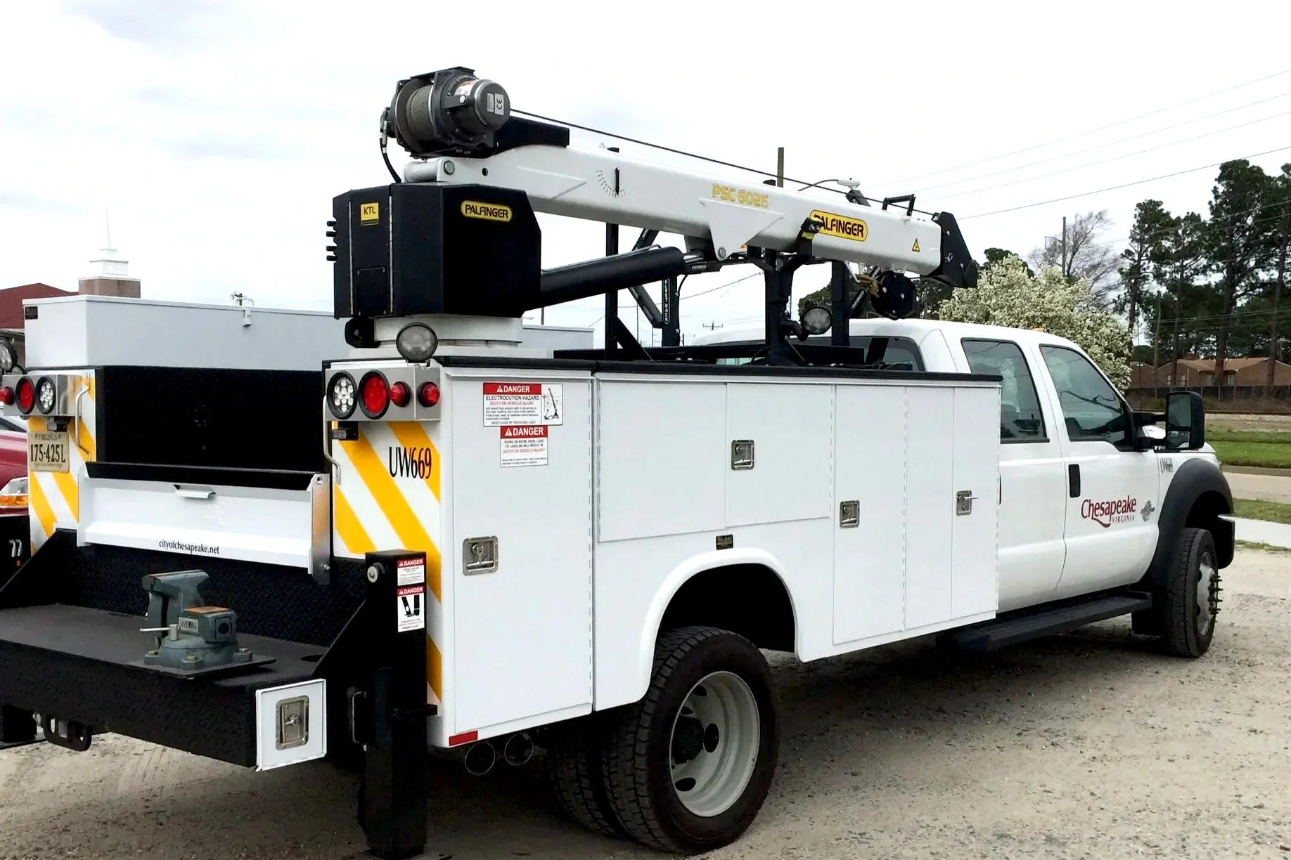 A service utility truck with a bucket lift and storage compartments, labeled with safety and city regulations, parked outdoors with trees and residential buildings in the background.