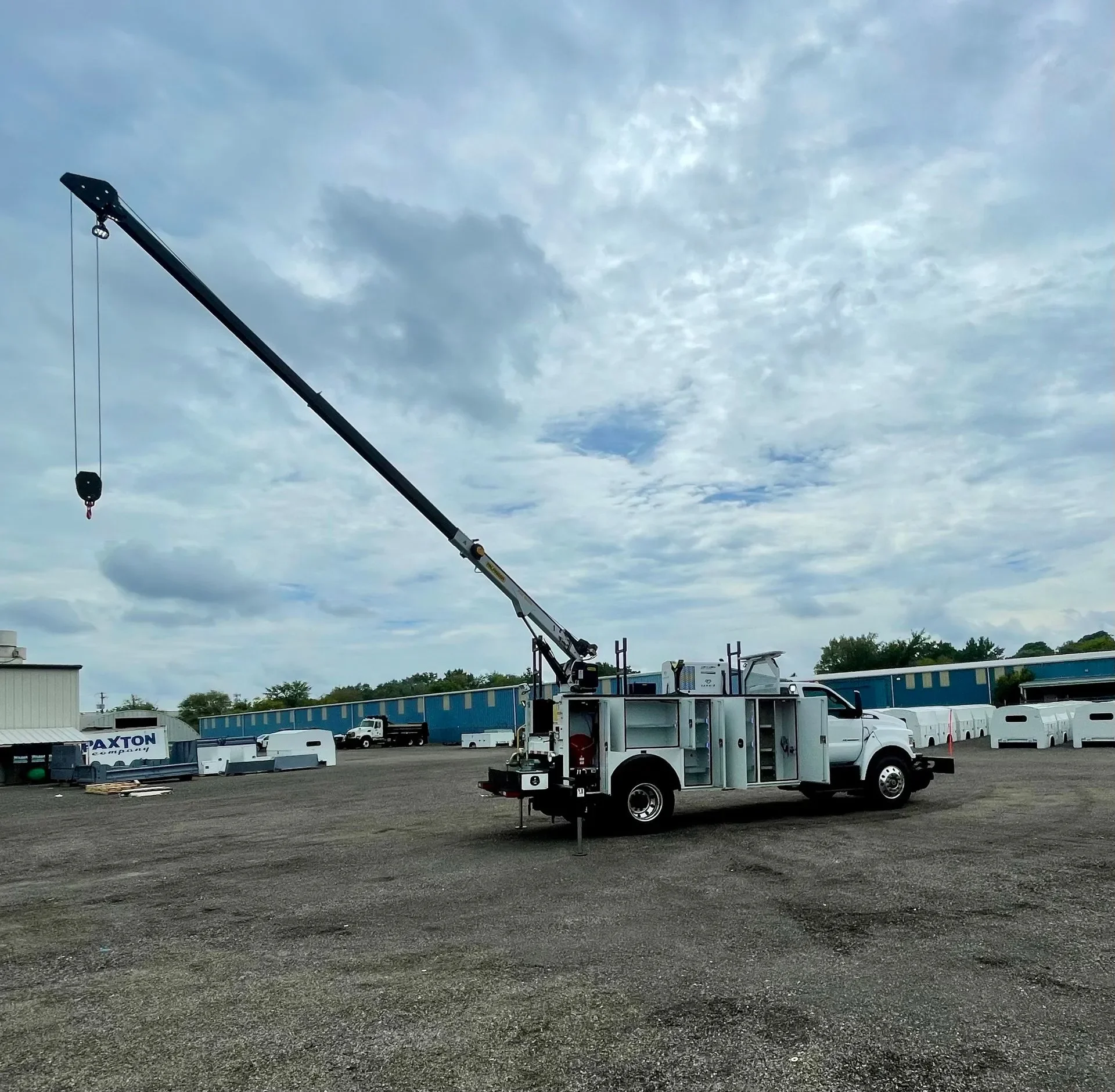 A mobile crane with a long black arm extended into the sky, standing on a gravel lot with trucks and blue industrial buildings in the background, under a cloudy sky.