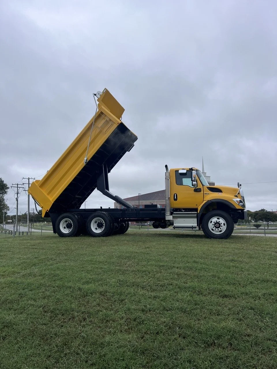 Yellow dump truck with raised bed parked on grass, overcast sky, residential area with buildings and power lines in the background.