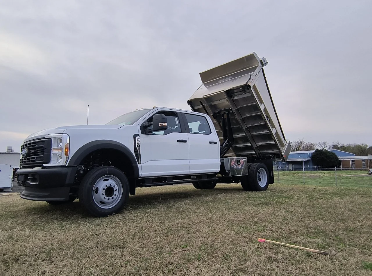 A white dump truck with its bed raised, parked on grass with a cloudy sky above.