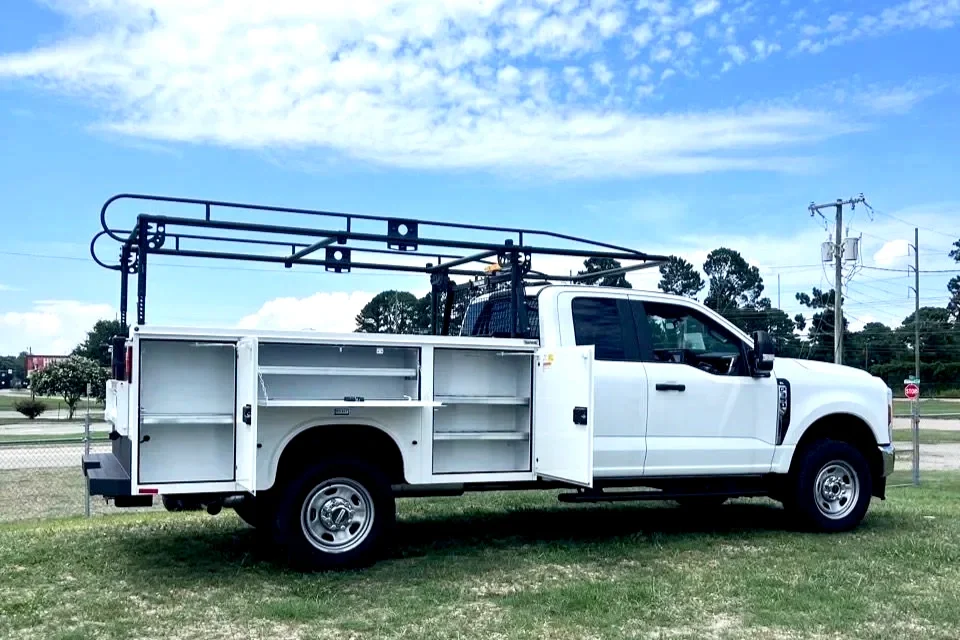 White utility truck equipped with a metal ladder rack and multiple storage compartments, parked on grass with a blue sky and power lines in the background.
