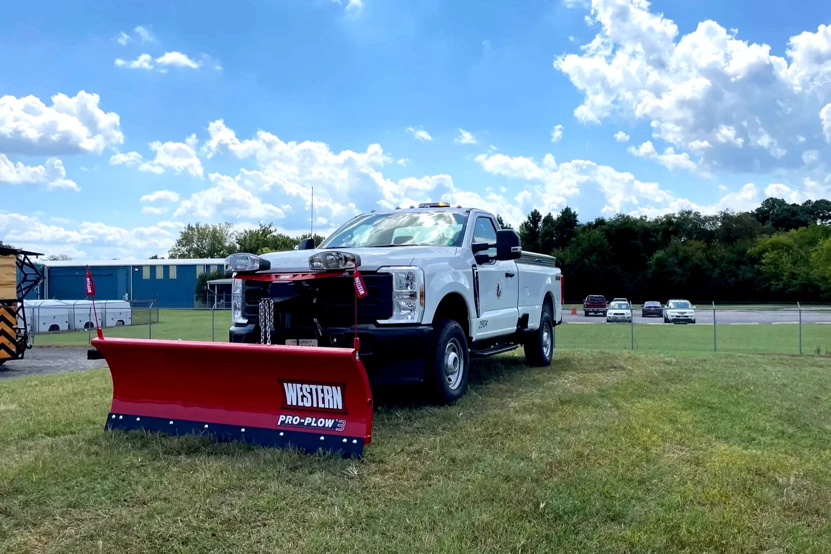 A white pickup truck with a large red snow plow attached to the front, parked on a grassy field on a sunny day with a few scattered clouds. There are several cars parked in the background and a chain-link fence surrounding the area.