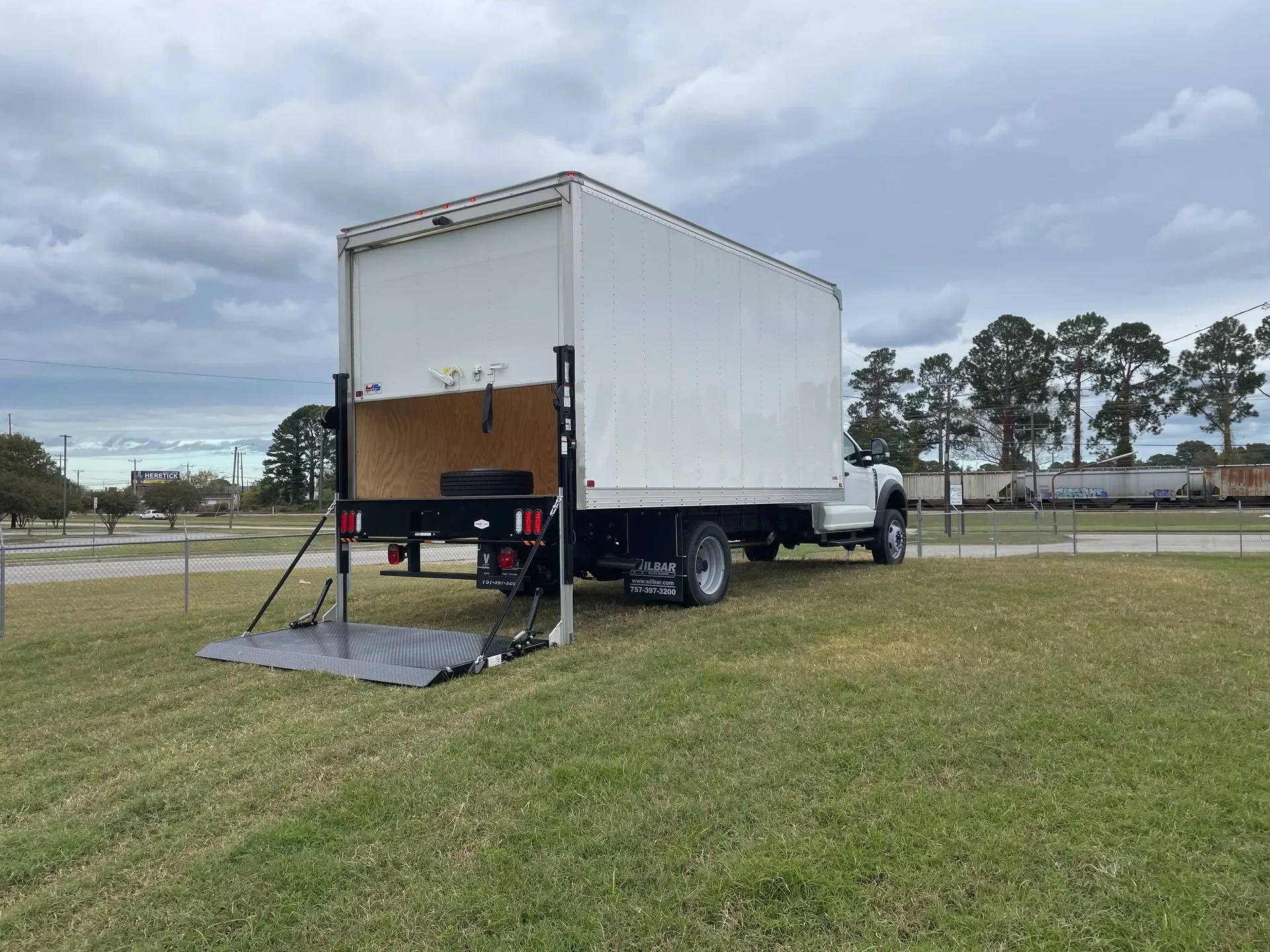 White moving truck with a wheelchair lift extended at a grassy area near a road, with trees and train cars in the background under a cloudy sky.