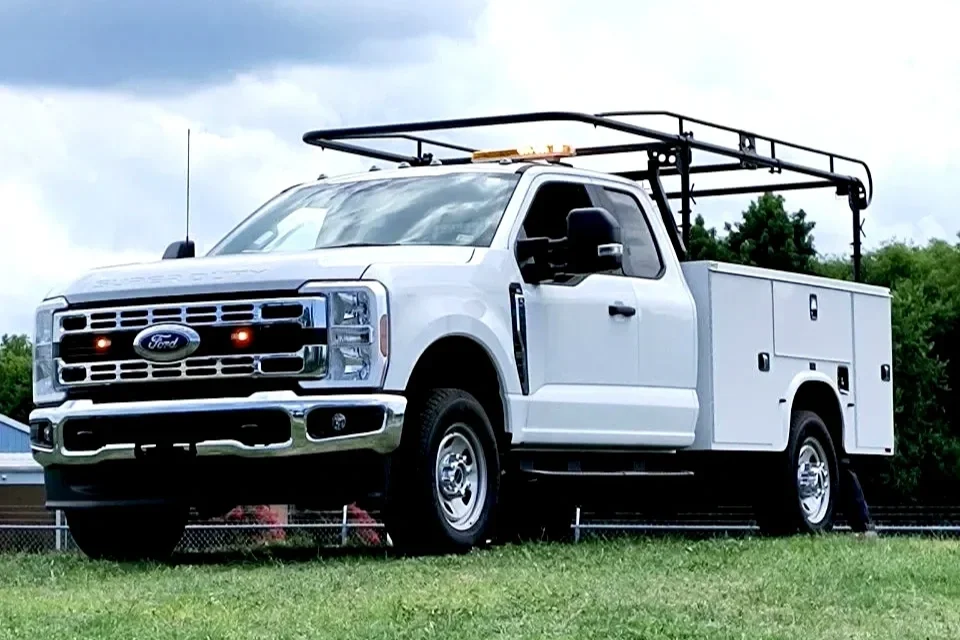 White Ford utility truck with a ladder rack on a grassy area, with trees and a cloudy sky in the background.