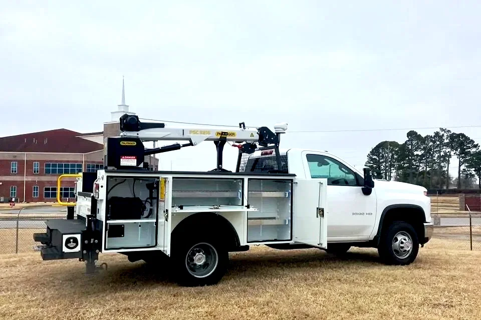 White utility truck with equipment storage and a crane on top, parked on a grassy field with a school building and trees in the background.