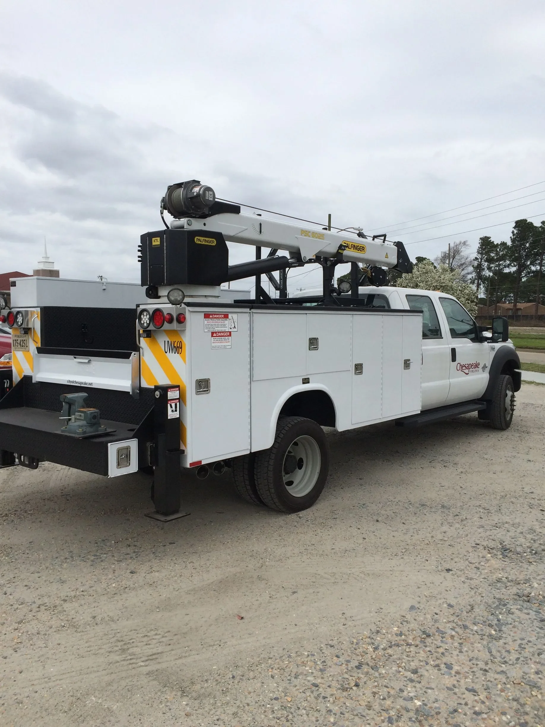 A utility bucket truck with an extendable boom and bucket, parked on a gravel lot under overcast skies, with trees and power lines in the background.