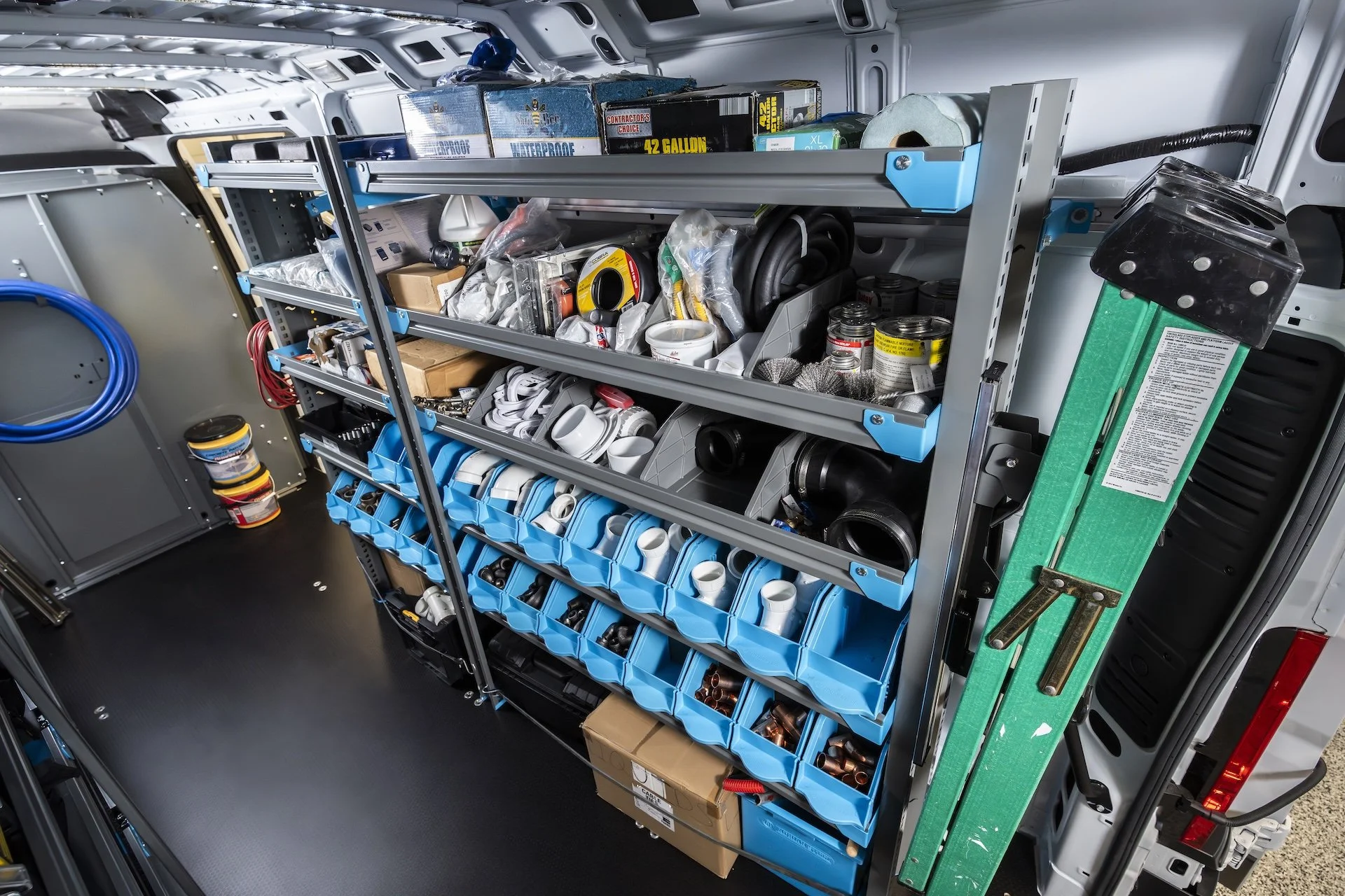 Interior view of a work van showing organized shelves filled with various tools, hardware, and supplies, including pipes, cans, and electrical components.