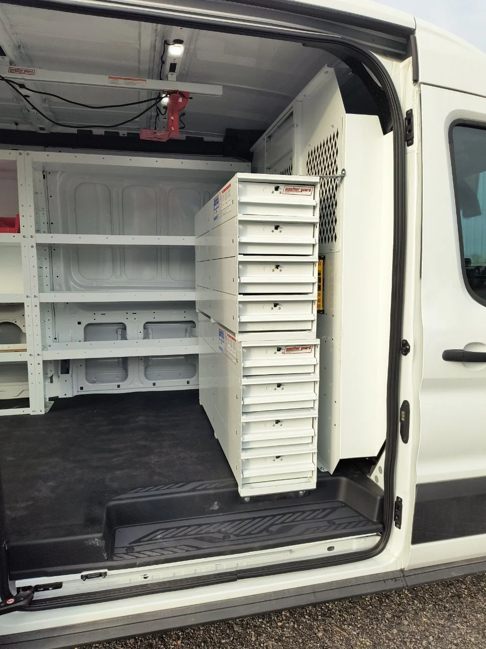 The interior of a white work van equipped with shelves and drawers, with the rear door open showing a black rubberized floor and organized storage units.