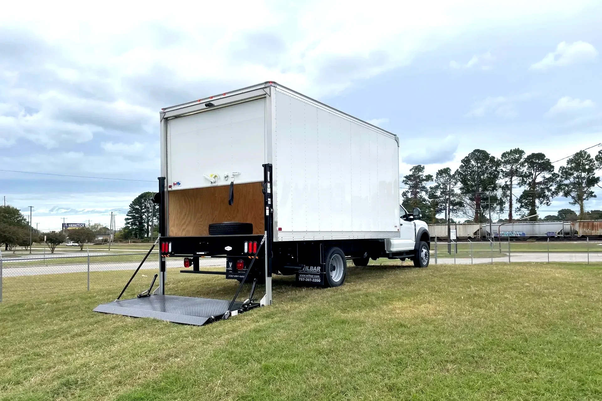 White moving truck with a tail lift and ramp extended, parked on grass near a fenced area with train tracks and trees in the background.