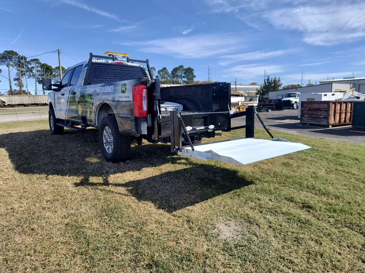 A John Deere vehicle with a lift gate and service branding parked on grass in an industrial area with trucks and storage containers in the background on a sunny day.