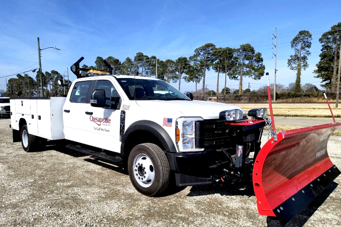 White utility truck with a red snowplow attachment in a parking lot, Chesapeake logos on the side, trees in the background, clear blue sky.