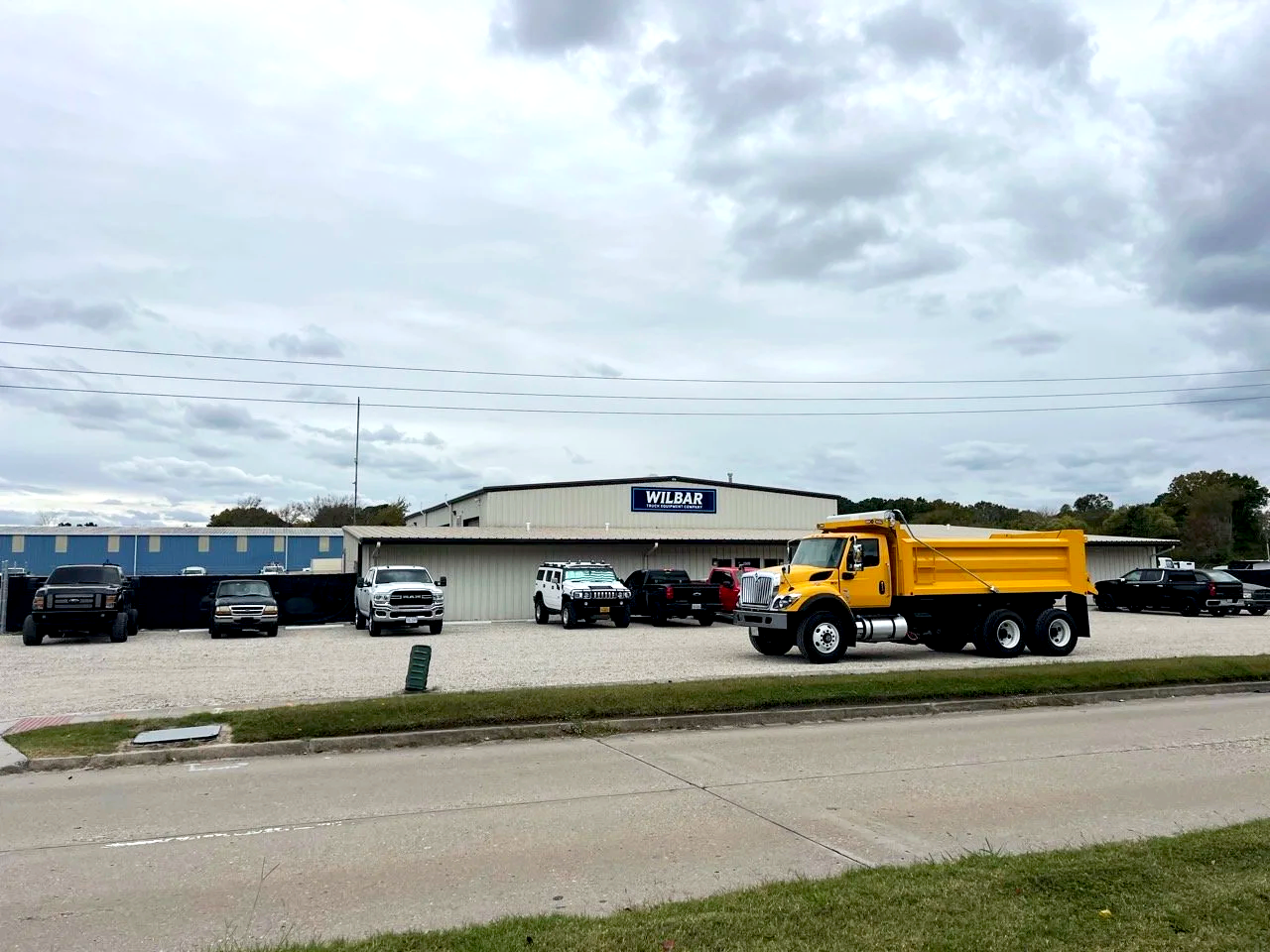 Parking lot in front of a building with a sign that reads 'Wilbar,' with several trucks and vehicles parked, including a yellow dump truck.