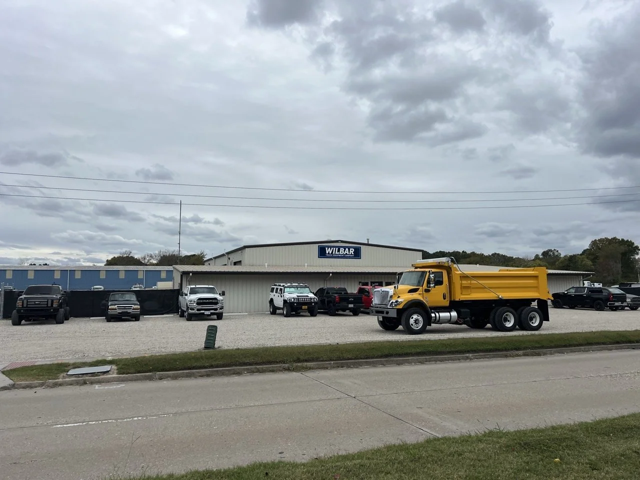 A commercial building with a sign that reads 'Wilbar' and a parking lot filled with trucks and SUVs. A yellow dump truck is parked in the foreground on the street.