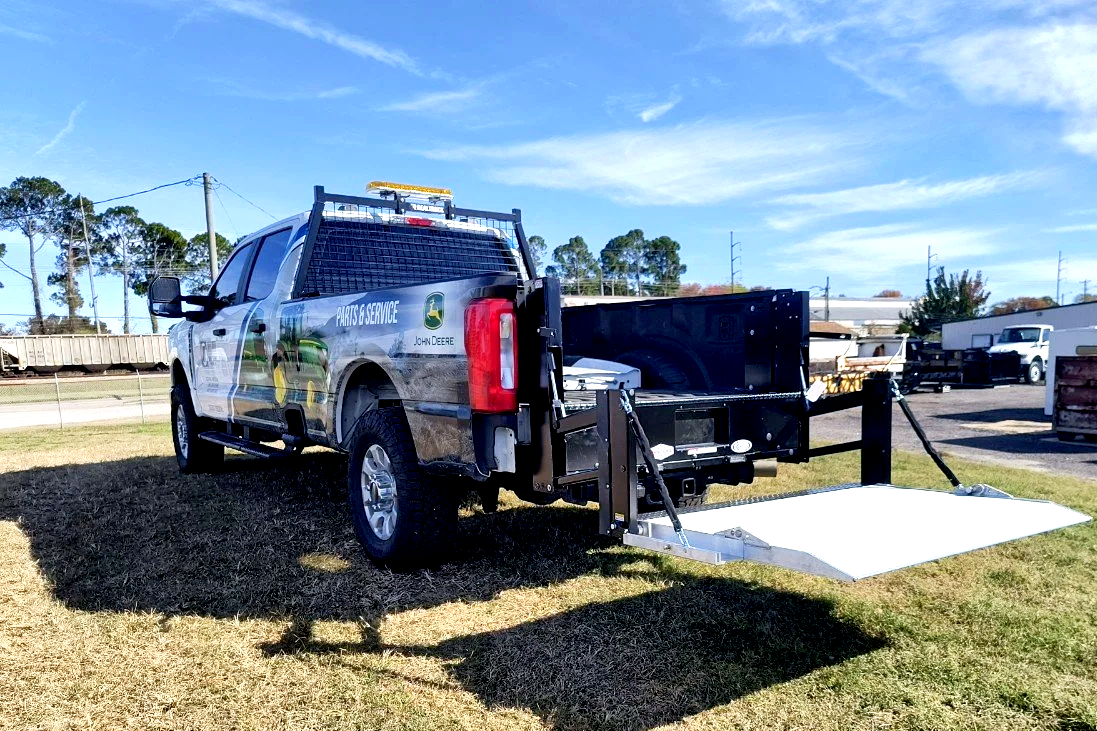A John Deere parts and service truck with a lift gate extended, parked on grass with other trucks and trees in the background.