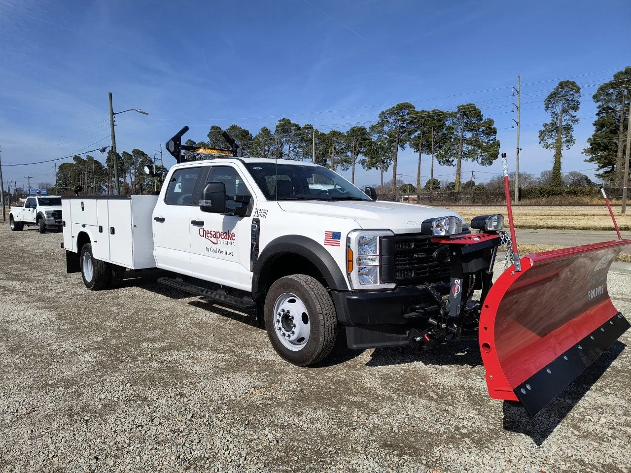 A white utility truck with a red snow plow attachment in a gravel lot, with trees and power lines in the background.