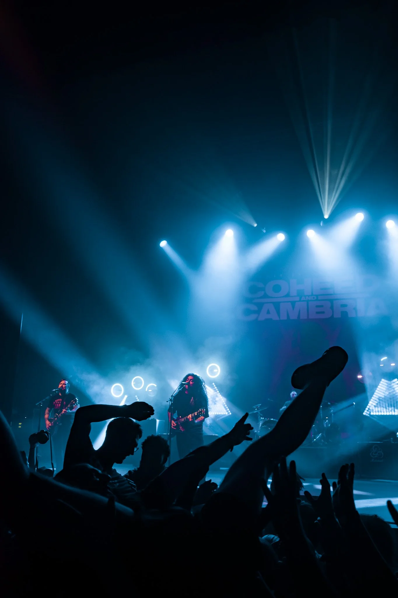 Concert stage with band performing under bright blue lights, silhouettes of audience members with raised hands and arms visible in the foreground.
