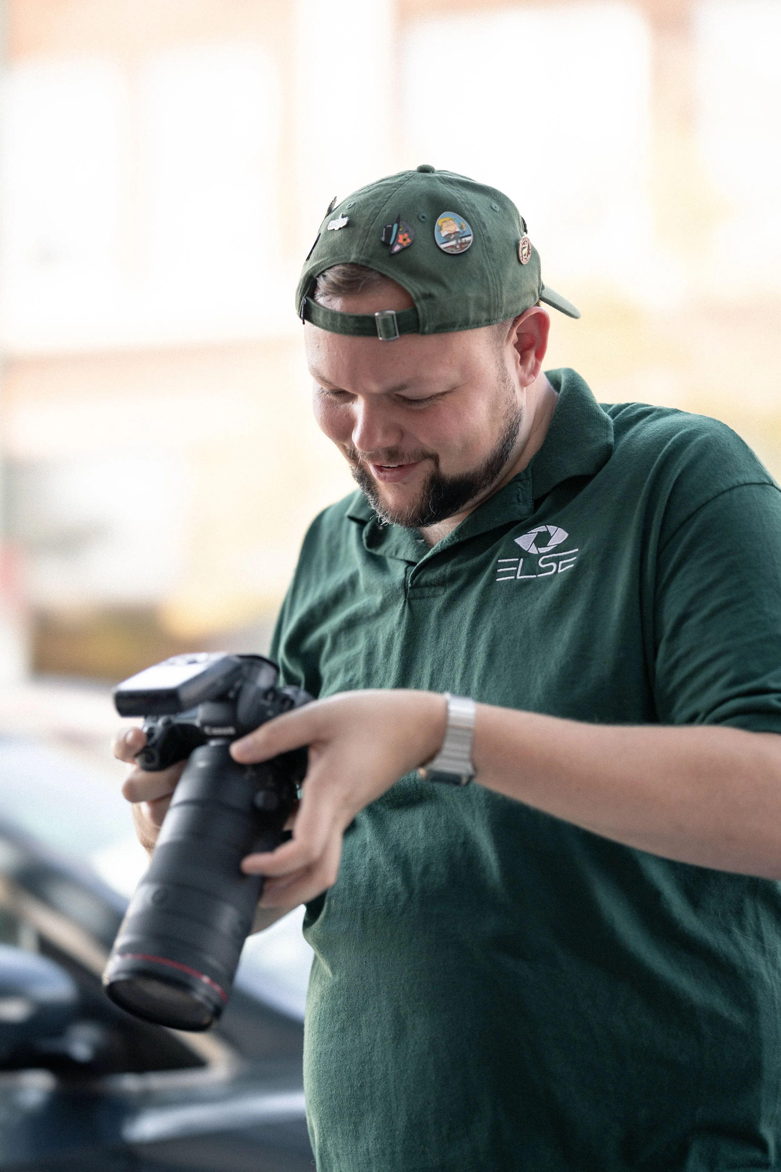 A man in a green polo shirt with a logo, wearing a green cap with various pins, holding a camera lens and looking at a camera.