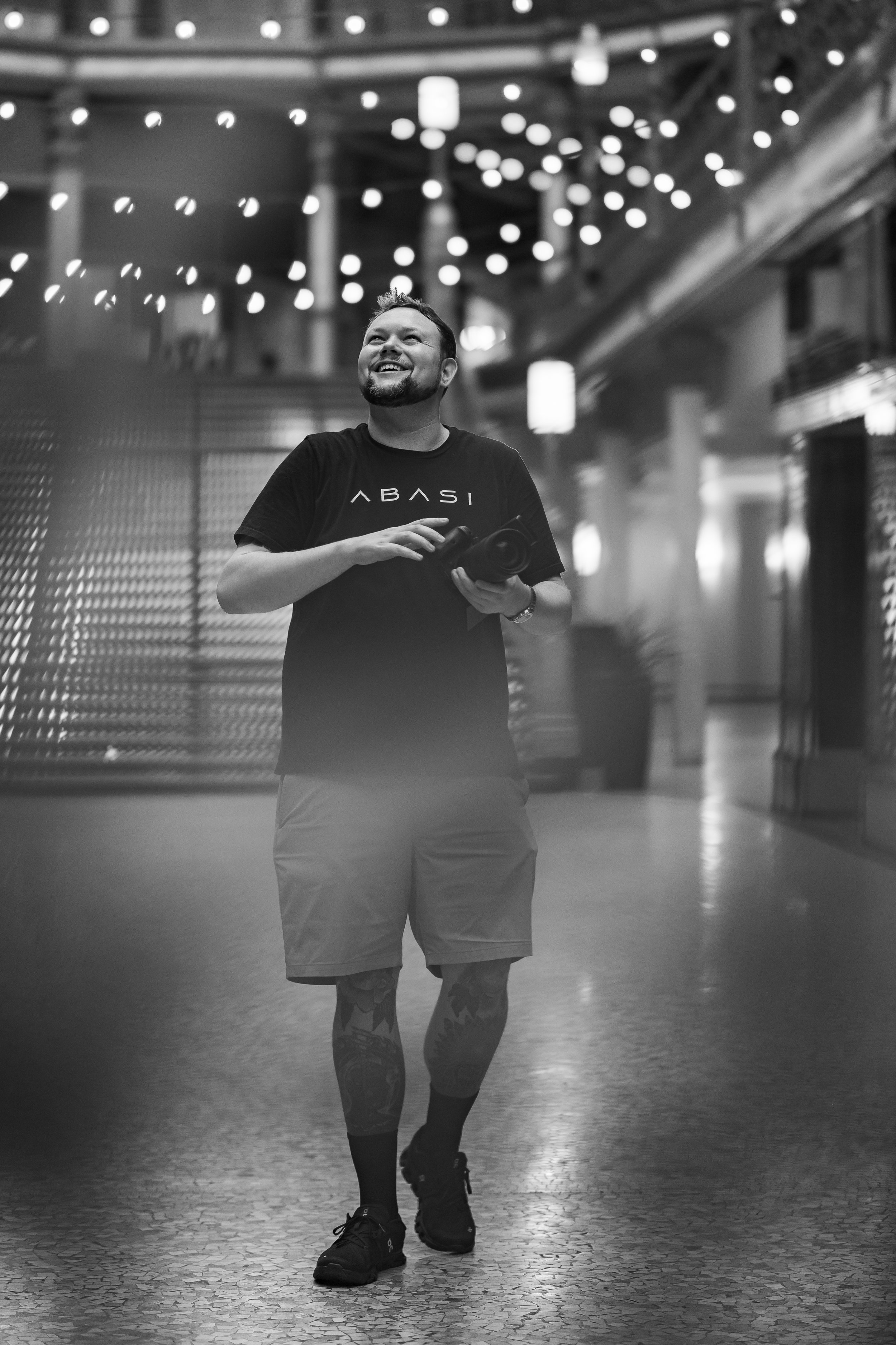 A man with a beard smiling and holding a camera, walking indoors with ceiling lights and modern decor, wearing a black t-shirt and shorts.