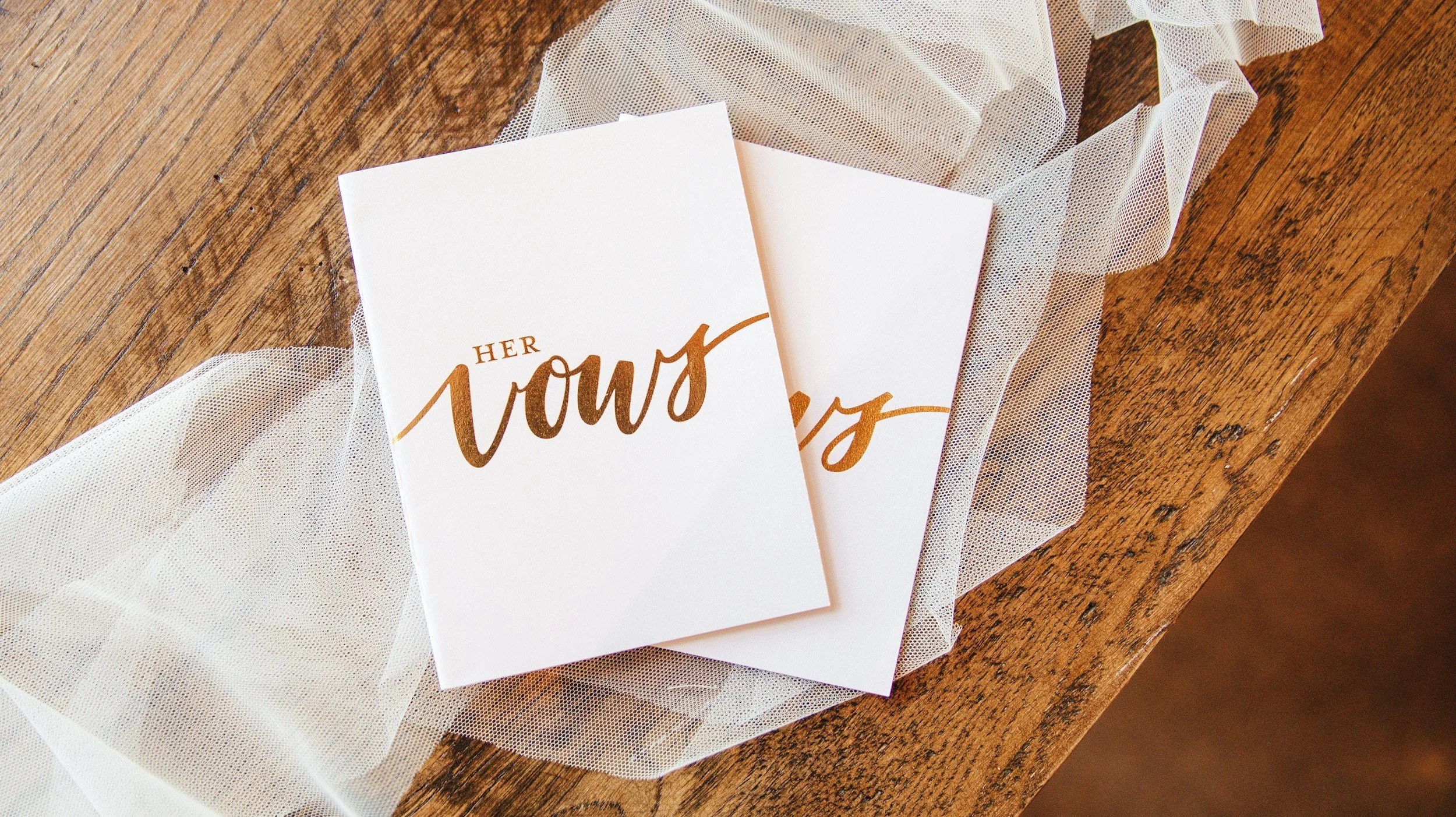 Wedding invitation with the words 'Her Vows' in gold script, placed on a wooden surface with a piece of white tulle fabric underneath.