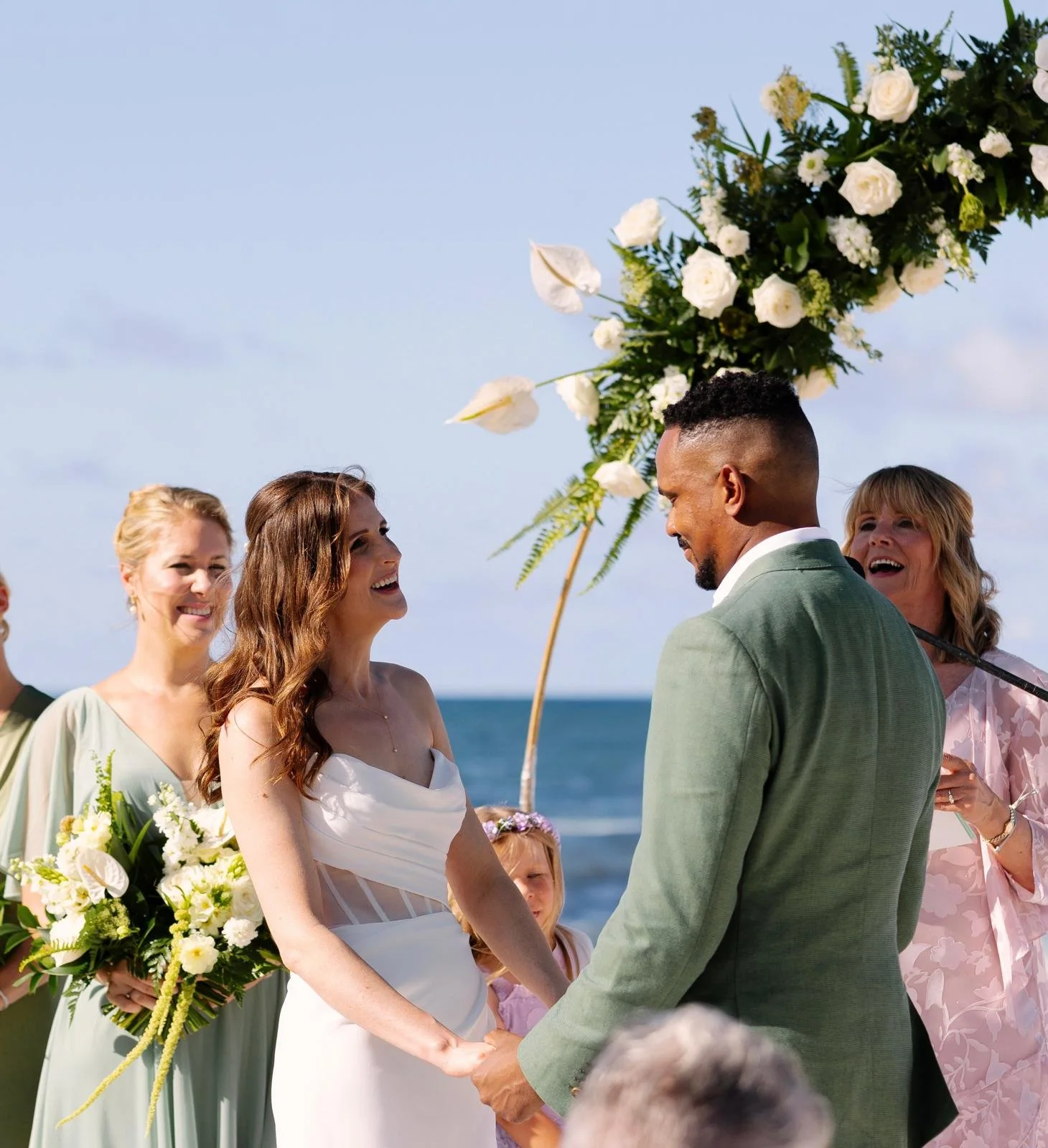 A wedding ceremony on the beach with a couple holding hands, surrounded by smiling women in pastel dresses, under a floral arch with white flowers and greenery, ocean in the background.