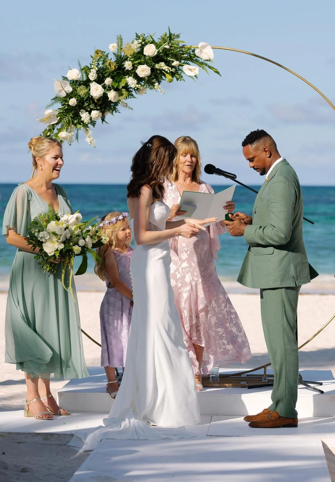 A beach wedding ceremony with a couple exchanging vows under a floral arch, surrounded by bridesmaids and a young girl, with an ocean background.