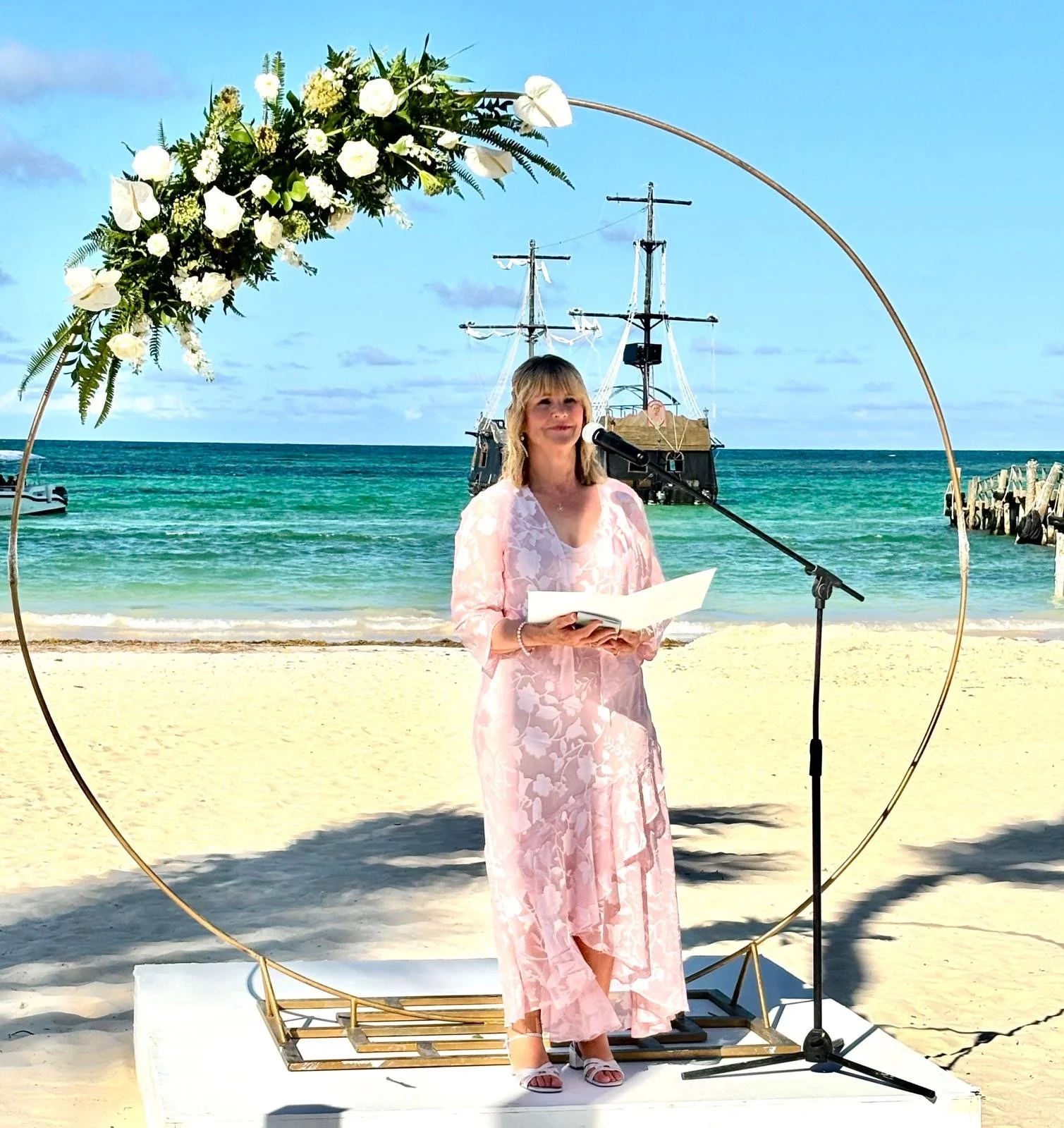 A woman in a pink floral dress standing on a beach, speaking into a microphone, with a ship in the background and a floral arch framing her.
