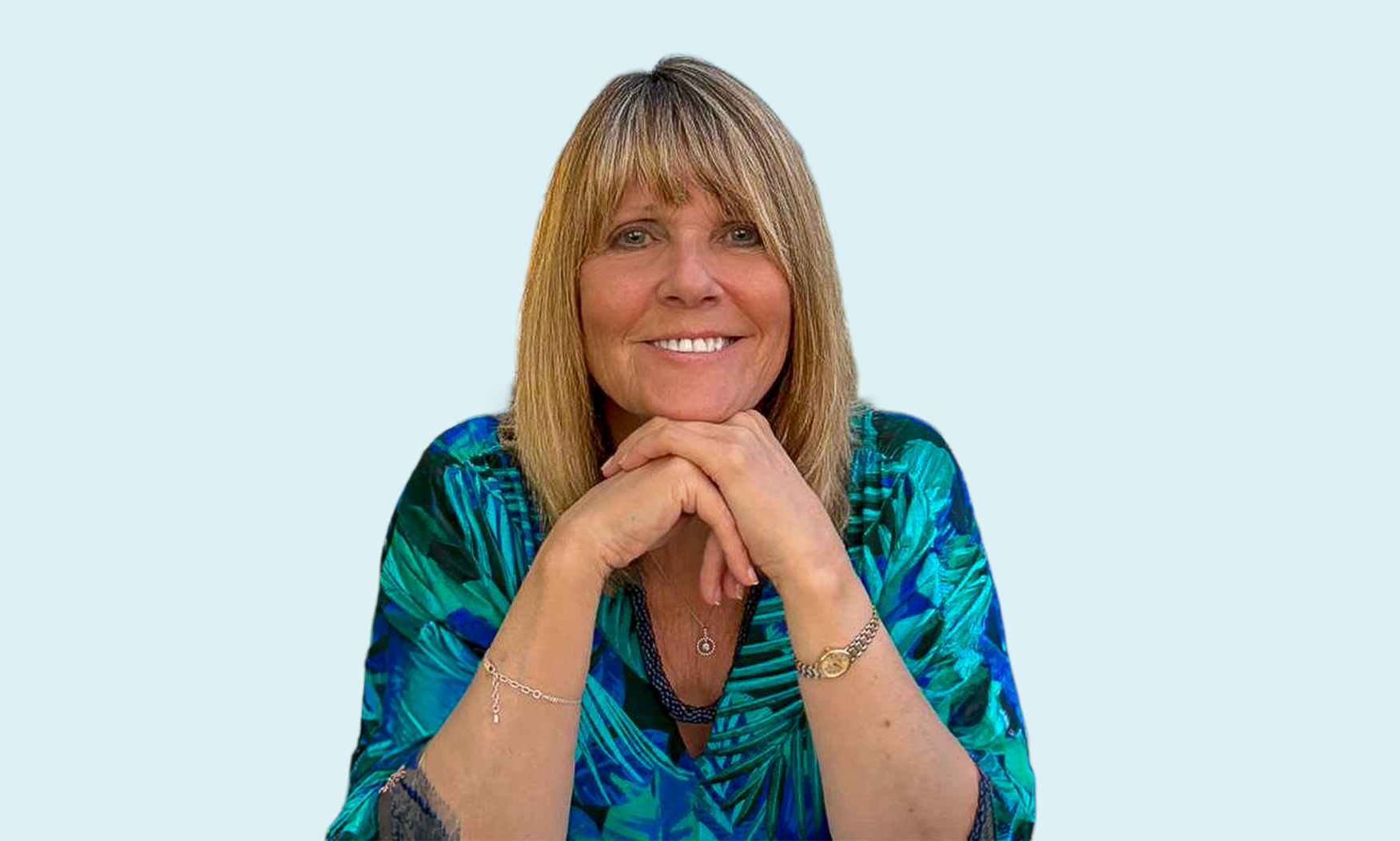 Annette Palmer with shoulder-length blonde hair smiling, resting her chin on her folded hands, wearing a blue and green patterned blouse, jewelry, and a silver necklace, against a light blue background.