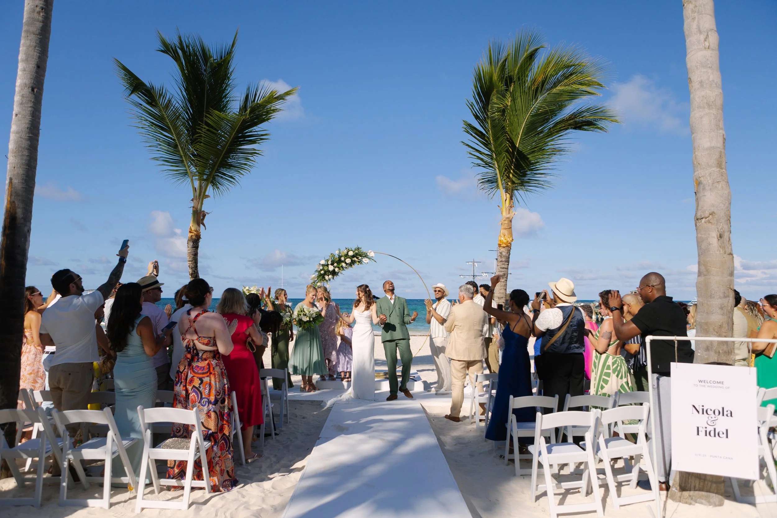 A beach wedding ceremony with a group of guests gathered around the officiant and couple at the altar, with palm trees and blue sky in the background.