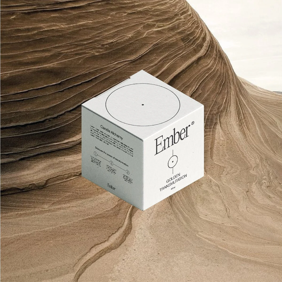 A white cube-shaped package labeled 'Ember' with text 'GOLDEN TRANSMUTATION' on a textured, sandy-colored surface with layered, eroded rock formations in the background.