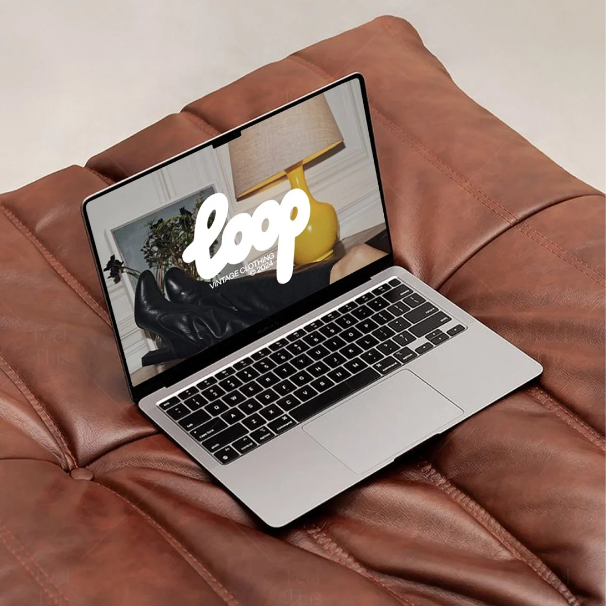 A laptop on a brown leather couch displaying a website for a vintage clothing brand called 'loop', with an image of black leather boots and a yellow table lamp in the background.