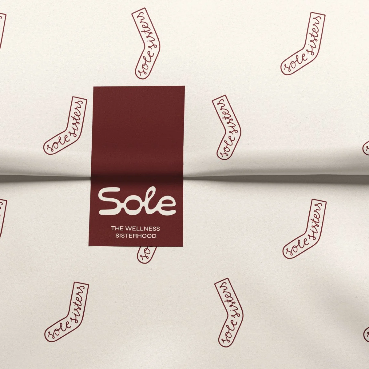 Close-up of wrapping paper with the word 'sole' and the phrase 'The Wellness Sisterhood' on a burgundy label, surrounded by repeating sock illustrations with the word 'sorefeeters' in red.
