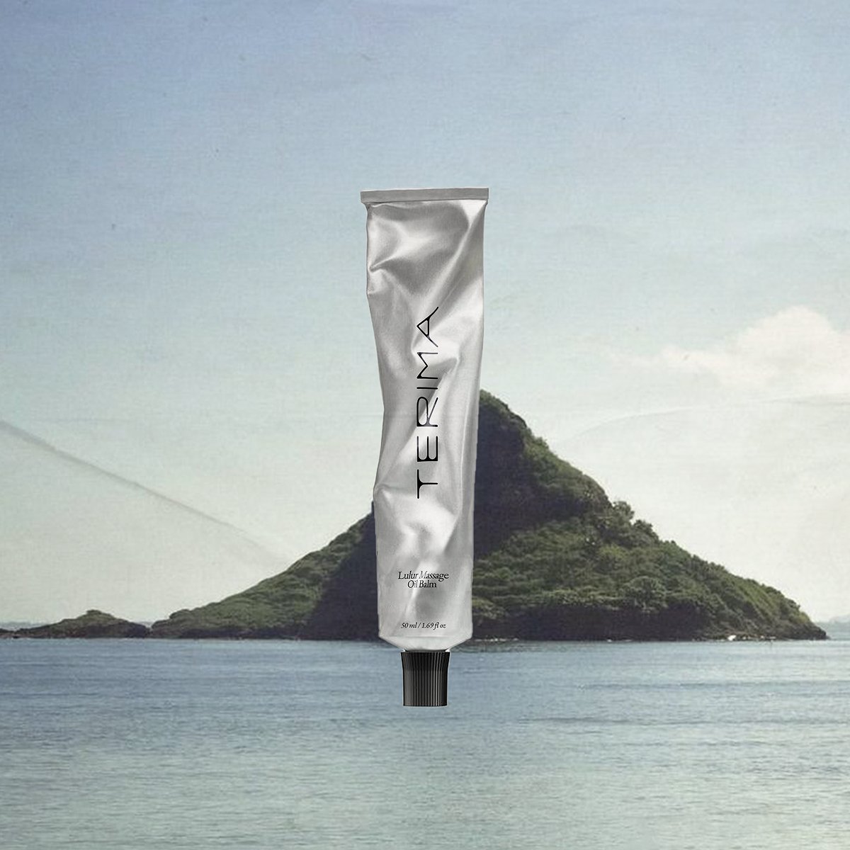 A silver tube of TEERIMA Lulur Massage Oil Balm floating in front of a scenic island with a mountain and water background.