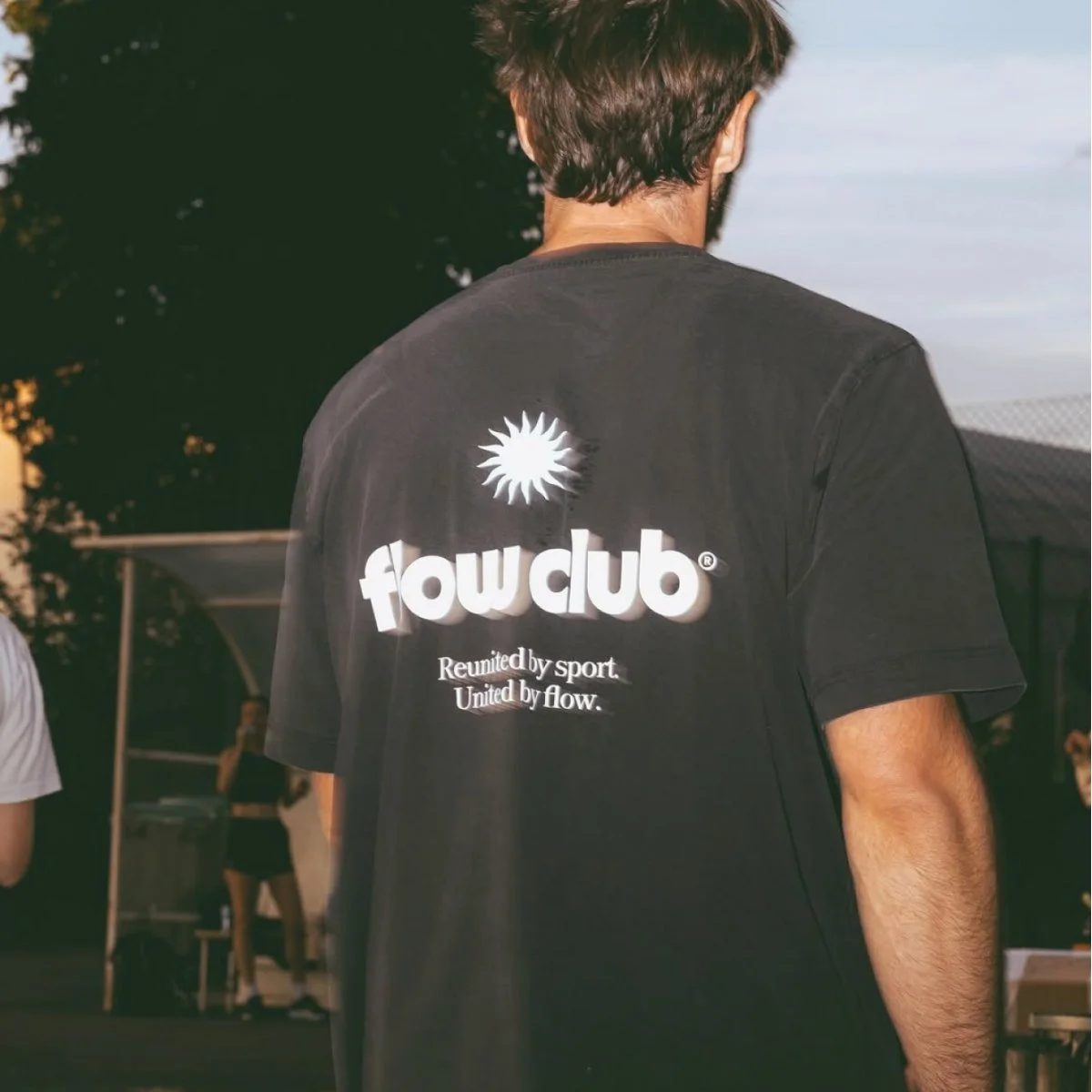 Back of a person wearing a black t-shirt with a logo and text, standing outdoors during sunset, with other people and trees visible in the background.