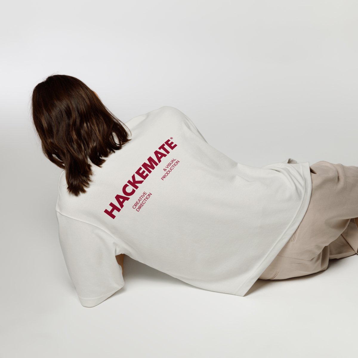 Person lying on their side with brown hair, wearing a white sweatshirt with red text that reads "HACKEMATE" and smaller text underneath that says "CREATIVE DIRECTION & VISUAL PRODUCTION."