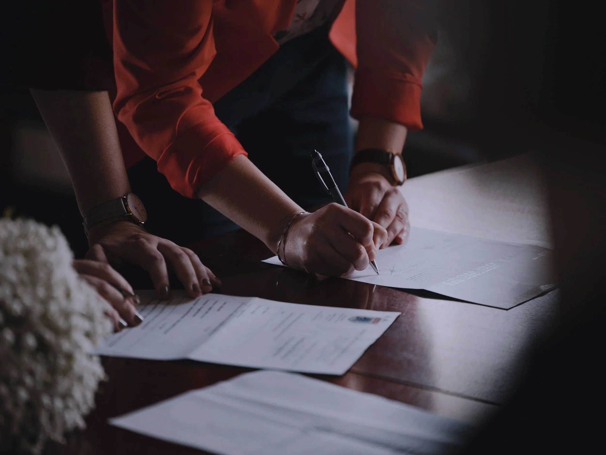 Person writing on paper with multiple sheets on a wooden table, wearing a red shirt and a watch.