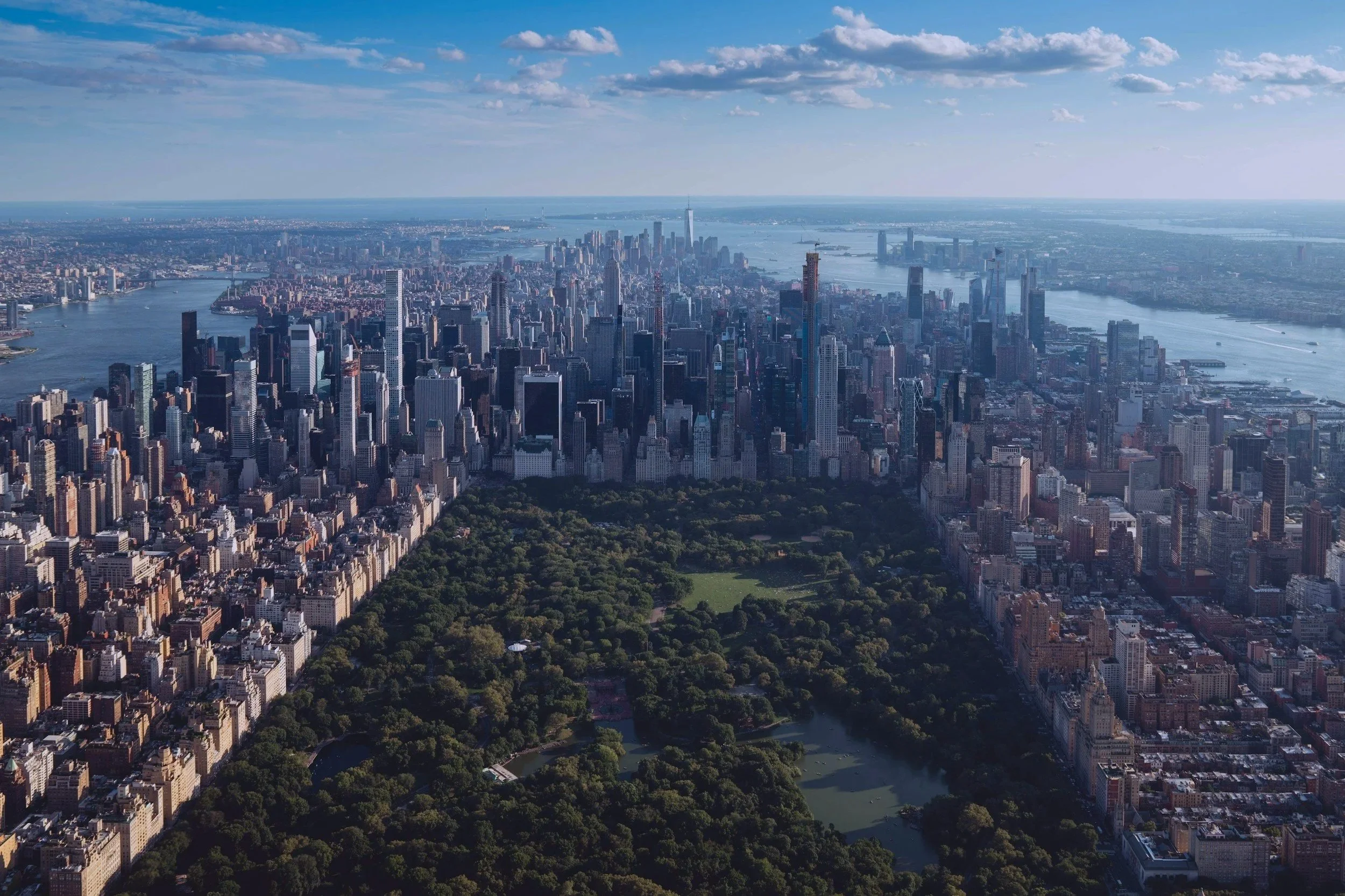 An aerial view of New York City skyline featuring tall skyscrapers, Central Park at the bottom, and the Hudson and East Rivers on either side, with a partly cloudy sky overhead.