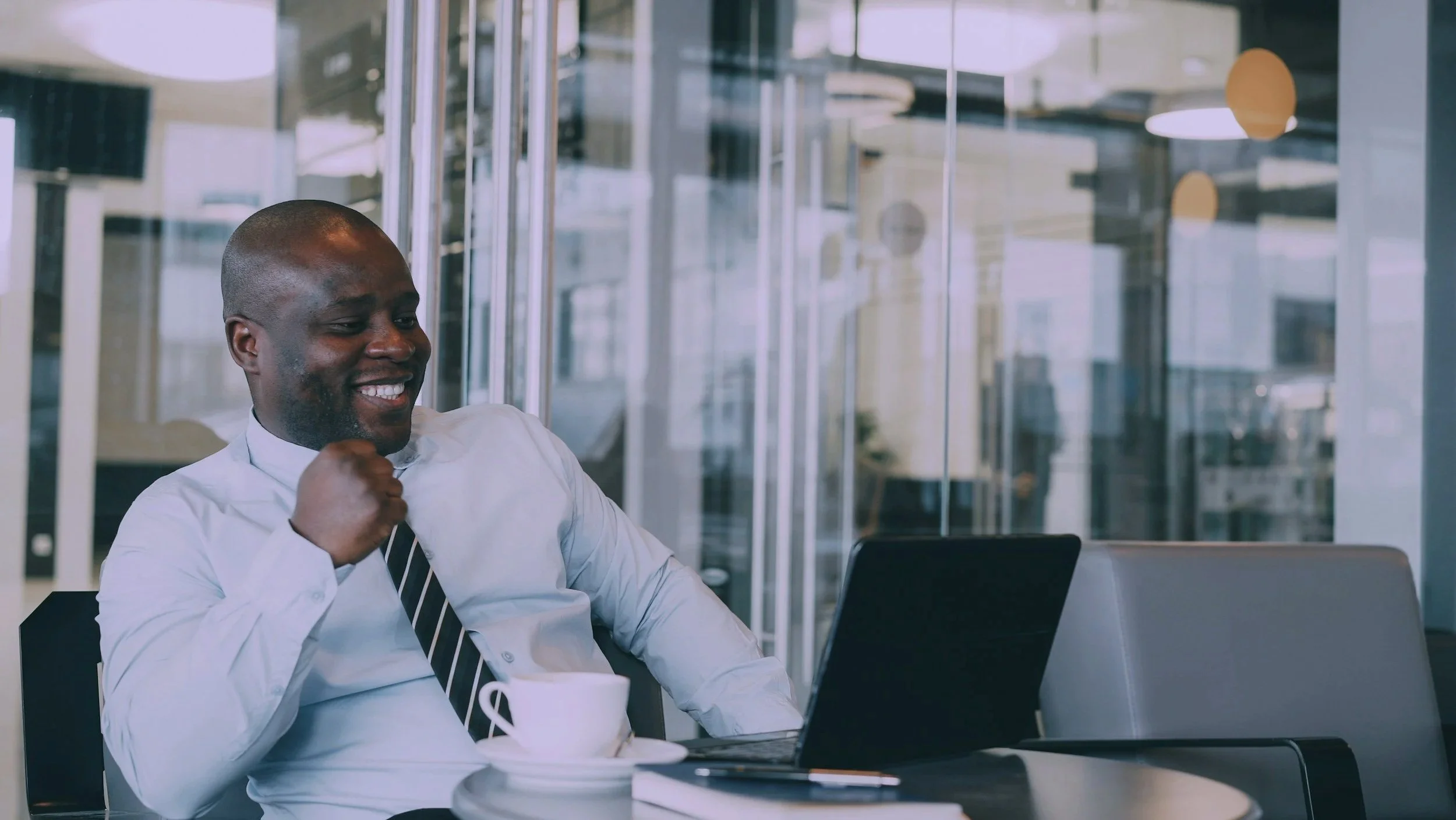 A smiling man in a white shirt and striped tie sitting at a desk in an office, with a laptop, cup, and notebook in front of him, in an office with glass walls.