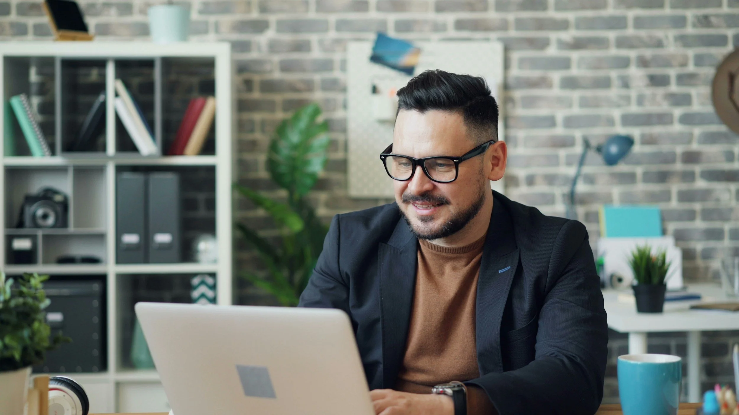 A man wearing glasses and a black blazer sitting at a desk in a modern office, looking at a laptop screen surrounded by office supplies and decor.
