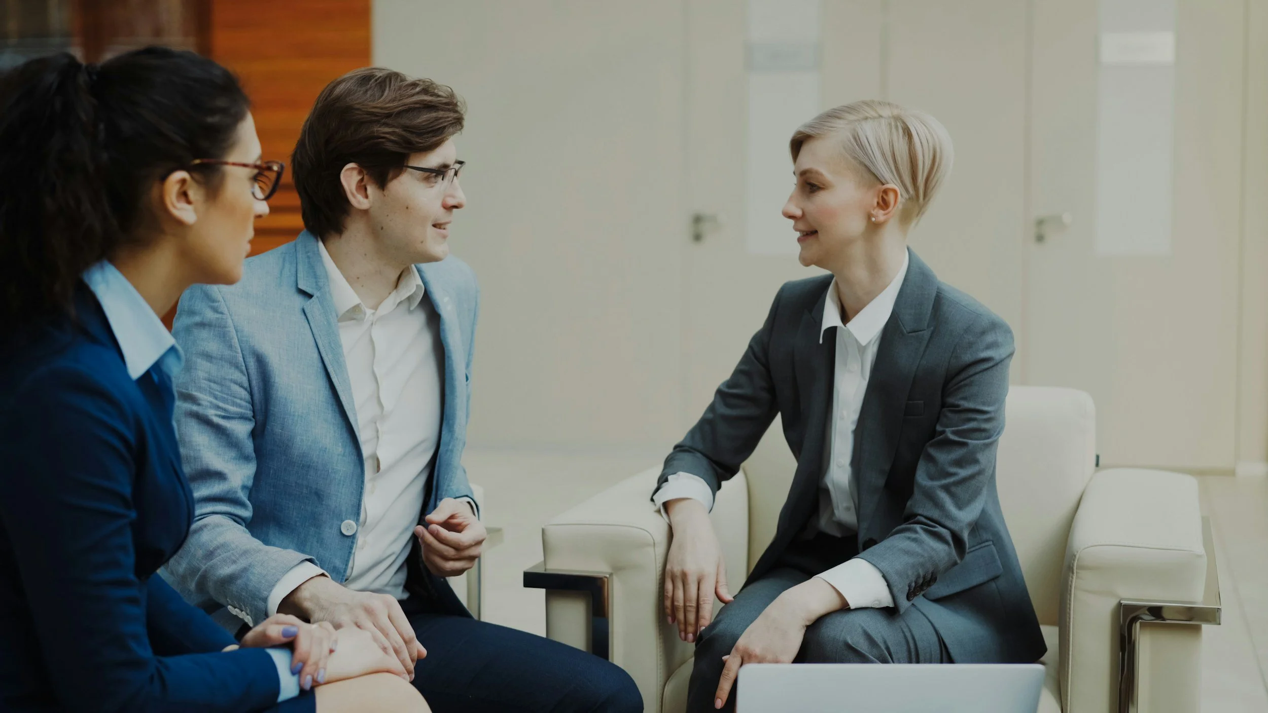 Four professional women are seated around a table engaging in conversation in a modern office setting.