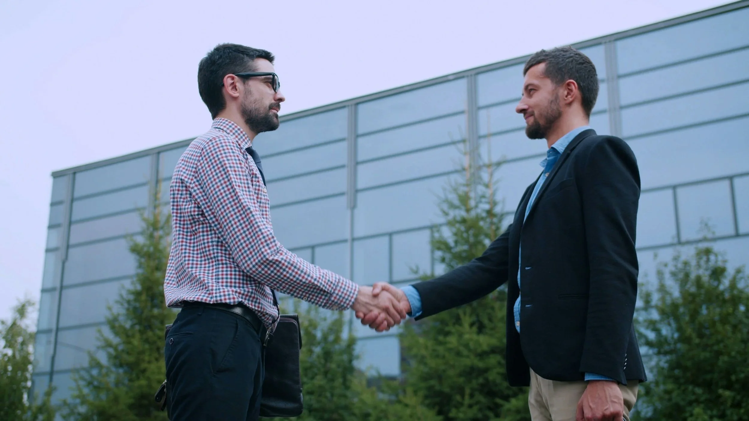 Two men shake hands outdoors in front of a modern building, dressed in business attire, with trees and a cloudy sky in the background.