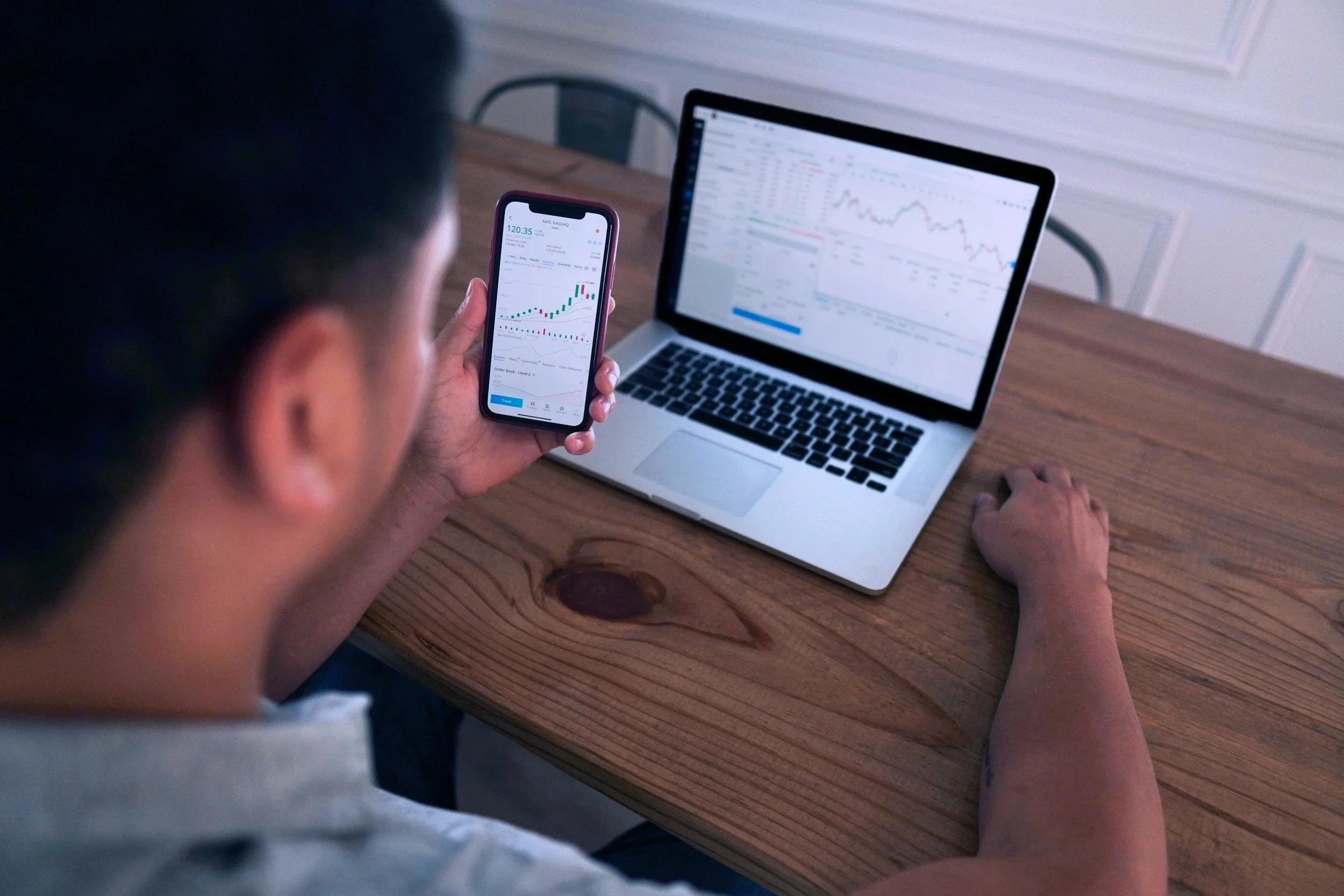 Person holding a smartphone displaying stock market graphs and data at a wooden desk with a laptop showing stock charts.