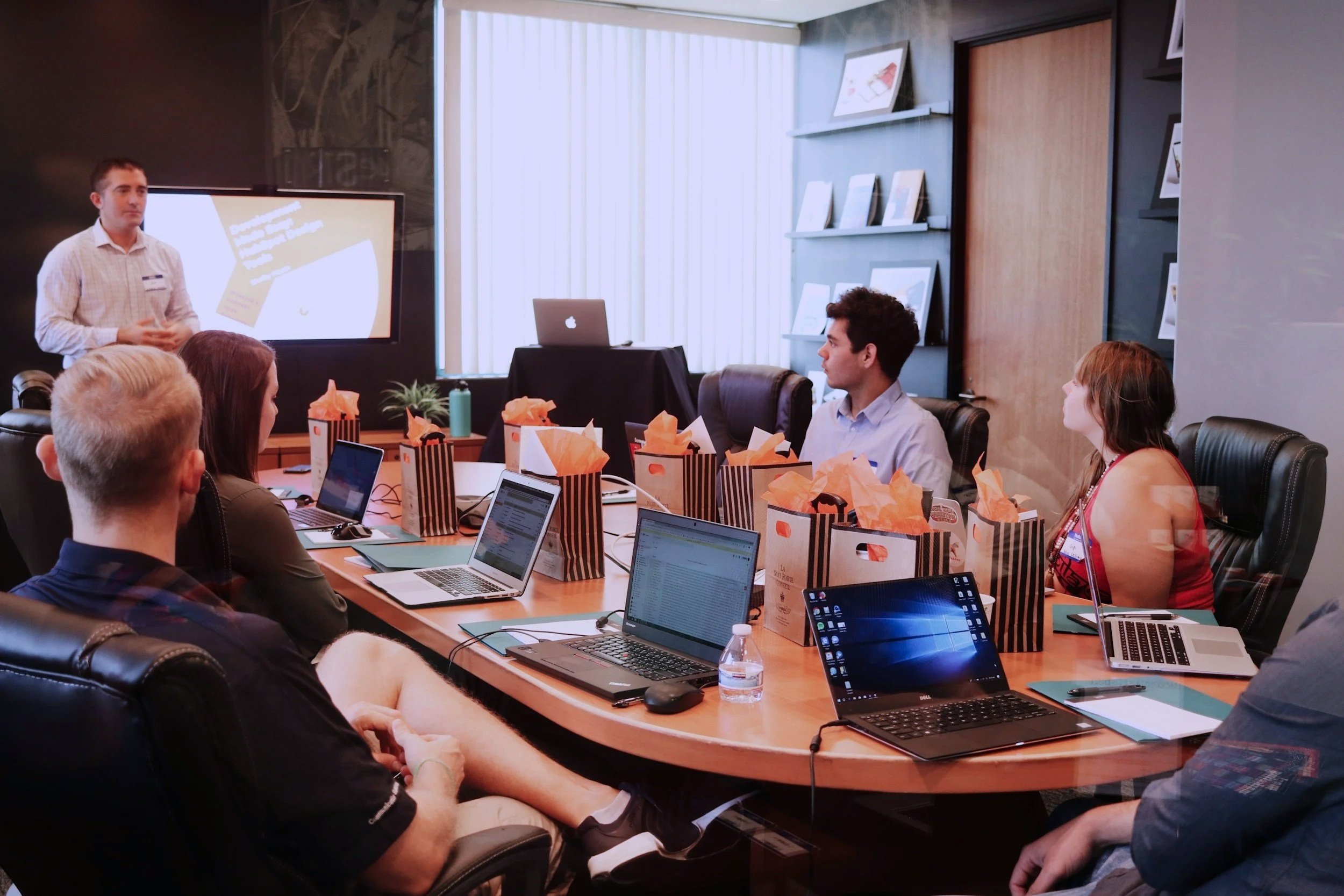 A group of people attending a business presentation or meeting in a conference room, with a man standing and speaking near a large screen, and several laptops and gift bags on the table.